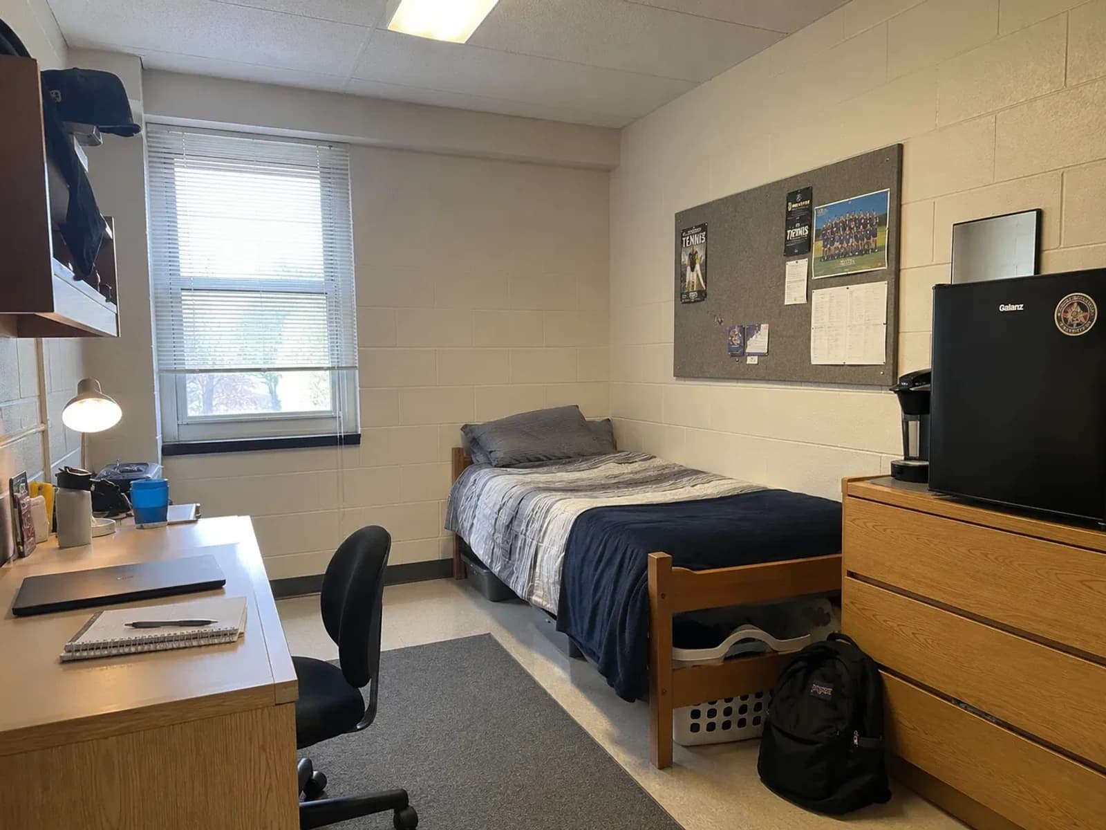 Existing small dorm room with cinder-block walls, raised bed, desk, shelves, dresser, compact refrigerator, and coffee maker