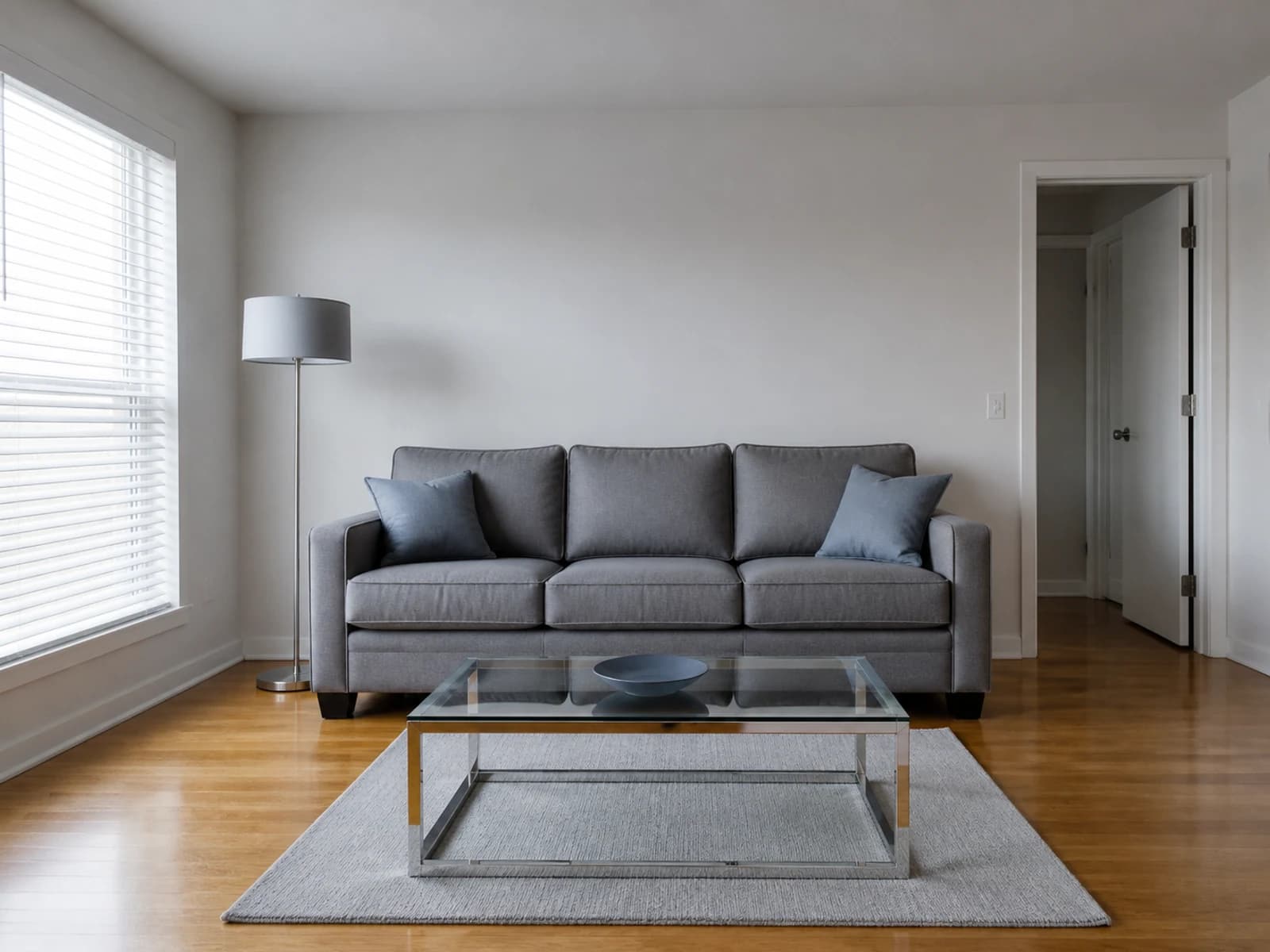 Cool unfinished living room with a medium-gray three-seat sofa, pale rug, glass table, and chrome lamp